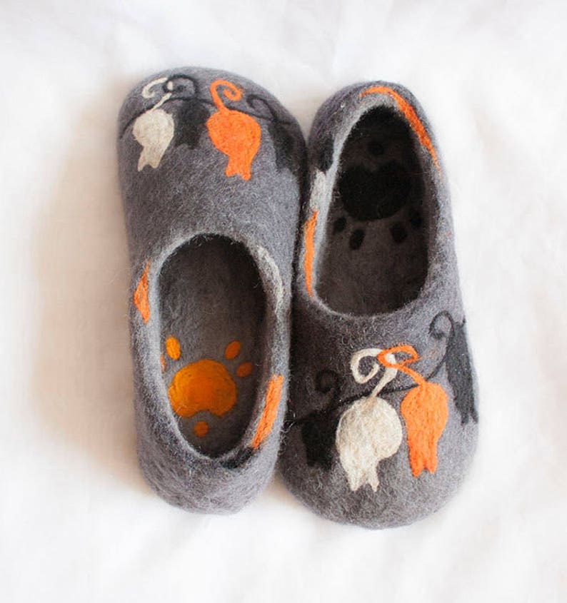 May include: A pair of gray felt slippers with orange, white, and black cat designs. The slippers have a paw print on the inside.