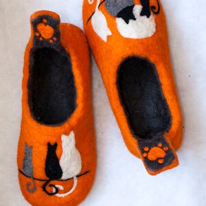May include: A pair of orange felt slippers with black and white cat designs. The slippers have a black felt lining and a paw print design on the heel.