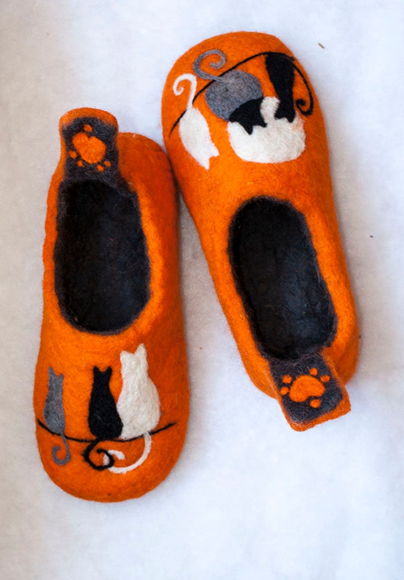 May include: A pair of orange felt slippers with black and white cat designs. The slippers have a black felt lining and a paw print design on the heel.