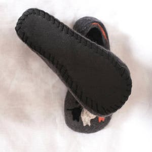 May include: A pair of gray wool slippers with a black leather sole. The slippers have a stitched edge and a design of a cat on the top.