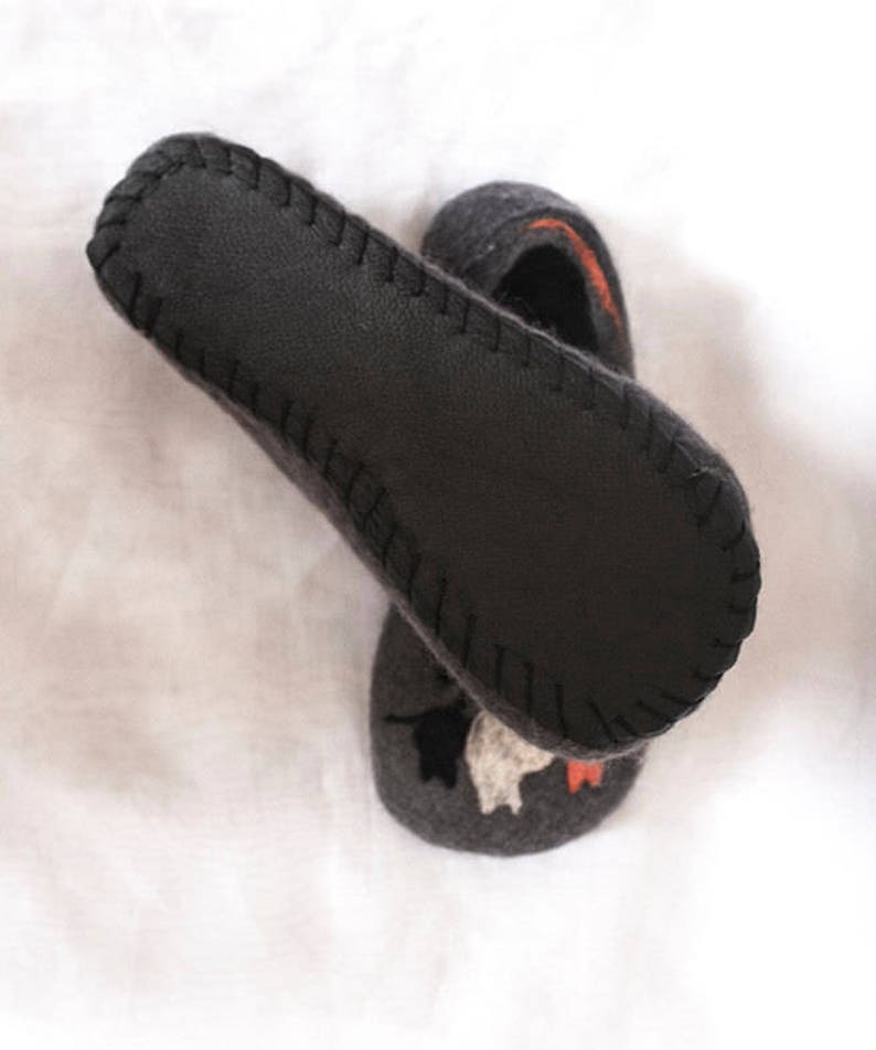 May include: A pair of gray wool slippers with a black leather sole. The slippers have a stitched edge and a design of a cat on the top.