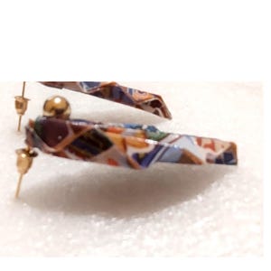 May include: A pair of earrings with a geometric pattern in shades of blue, brown, and orange. The earrings have a gold-colored post and a small gold ball. The earrings are on a white surface.