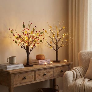 Op de afbeelding: Twee decoratieve verlichte boomsculpturen met bruine stammen en bases. Eén boom heeft rode bessen en groene bladeren, de andere heeft gouden bladeren. Beide hebben warmwitte lampjes. Ze staan op een houten tafel.