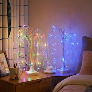 Può includere: Due lampade decorative a forma di salice piangente con luci in fibra ottica. Una lampada ha luci multicolori, l'altra luci blu. Le lampade hanno basi bianche e tronchi simili a betulle, creando un'atmosfera calda.