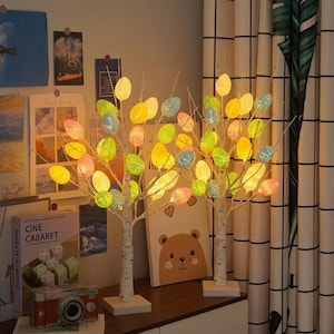 May include: Two decorative lighted birch trees with colorful egg-shaped lights. The lights are in shades of yellow, green, pink, and blue. The trees are on white square bases, and the background includes books, a camera, and a bear illustration.