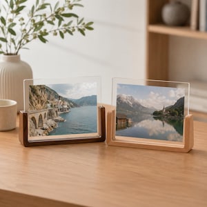 May include: Two rectangular acrylic photo frames with wooden bases. The left frame has a dark walnut base and displays a coastal landscape with cliffs and water. The right frame has a light oak base and shows a mountain lake scene with a small cabin.
