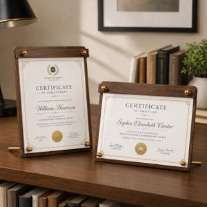 May include: Two dark wood document frames with gold-toned metal hardware. The vertical frame displays a Certificate of Achievement for William Harrison, while the horizontal frame displays a Certificate of Completion for Sophia Elizabeth Carter.