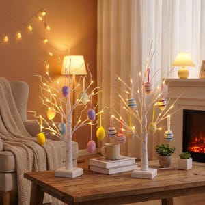 May include: Two white birch trees with LED lights and colorful Easter egg ornaments. The trees sit on a wooden table with a neutral-toned background. A cozy, festive decoration for the Easter holiday.