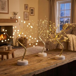 May include: Three LED tree lights with warm white lights. The trees have gold, silver, and gold finishes, with round or star-shaped lights. They are placed on a wooden table in a cozy living room setting.