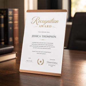 Puede incluir: Una placa de reconocimiento rectangular con una superficie blanca y texto en dorado y negro. El encabezado dice Recognition Award, seguido del nombre Jessica Thompson, una declaración formal, la fecha 15 de mayo de 2024 y una firma.