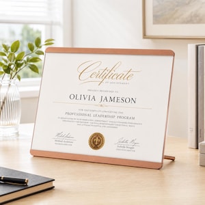 May include: A rectangular certificate of achievement in a metallic rose gold frame. The document features the text Certificate of Achievement, Olivia Jameson, Professional Leadership Program, and signatures from Michael Anderson and Isabella Morgan.