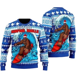 May include: A blue long-sleeve jumper featuring a graphic of a brown Bigfoot wearing a Santa hat and sunglasses while surfing on a red surfboard. The text BIGFOOT SURFING SWELLS is printed above the wave. The jumper has a patterned border.