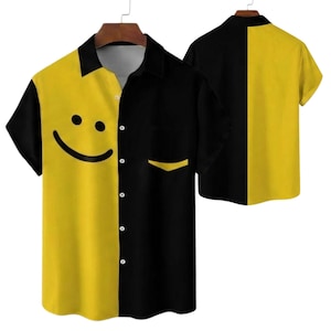 Puede incluir: Una camisa de manga corta con un diseño dividido. Una mitad es de color amarillo brillante con un gráfico de cara sonriente, y la otra mitad es negra. Un bolsillo está en el lado negro. La parte posterior de la camisa también es visible.