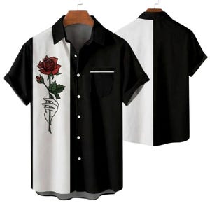 Puede incluir: Camisa de manga corta con un diseño de bloques de color blanco y negro. El lado izquierdo presenta un gráfico de una mano sosteniendo una rosa roja. La camisa tiene un bolsillo en el pecho izquierdo.