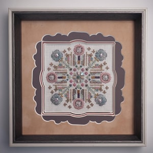 May include: Framed cross-stitch artwork featuring a symmetrical floral design in shades of pink, blue, and gold. The piece is embellished with beads and sequins, set against a neutral fabric. The artwork is framed in a dark wood frame with a light-colored inner border.