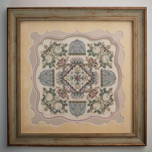 Puede incluir: Obra de arte en punto de cruz enmarcada con un patrón central de diamante rodeado de diseños florales y geométricos. La pieza está en un marco dorado envejecido con un paspartú beige claro y un borde decorativo en tonos rosa y beige.