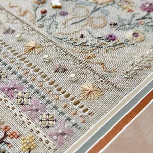 May include: A framed embroidery sampler on light beige linen fabric featuring intricate floral motifs, geometric patterns, and decorative drawn thread work. The design includes pastel-colored thread, small beads, and a white butterfly embellishment.