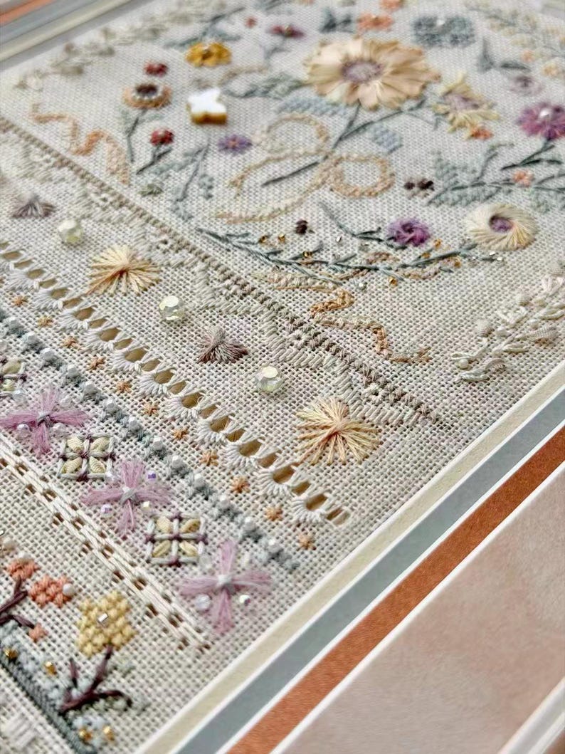 May include: A framed embroidery sampler on light beige linen fabric featuring intricate floral motifs, geometric patterns, and decorative drawn thread work. The design includes pastel-colored thread, small beads, and a white butterfly embellishment.