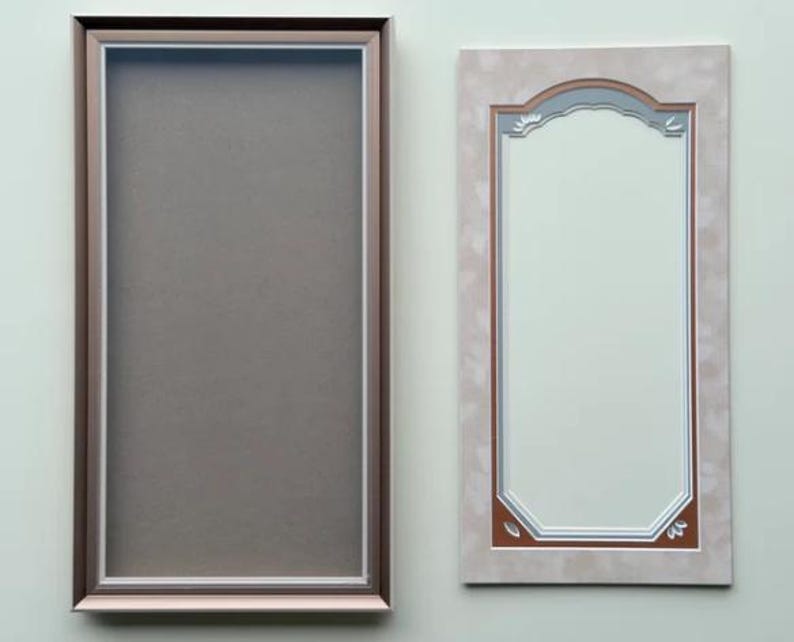 May include: A rectangular frame with a metallic bronze finish and a white inner border surrounding a solid gray center panel, positioned next to a rectangular mirror with a light beige patterned frame and a decorative arched top edge detail.
