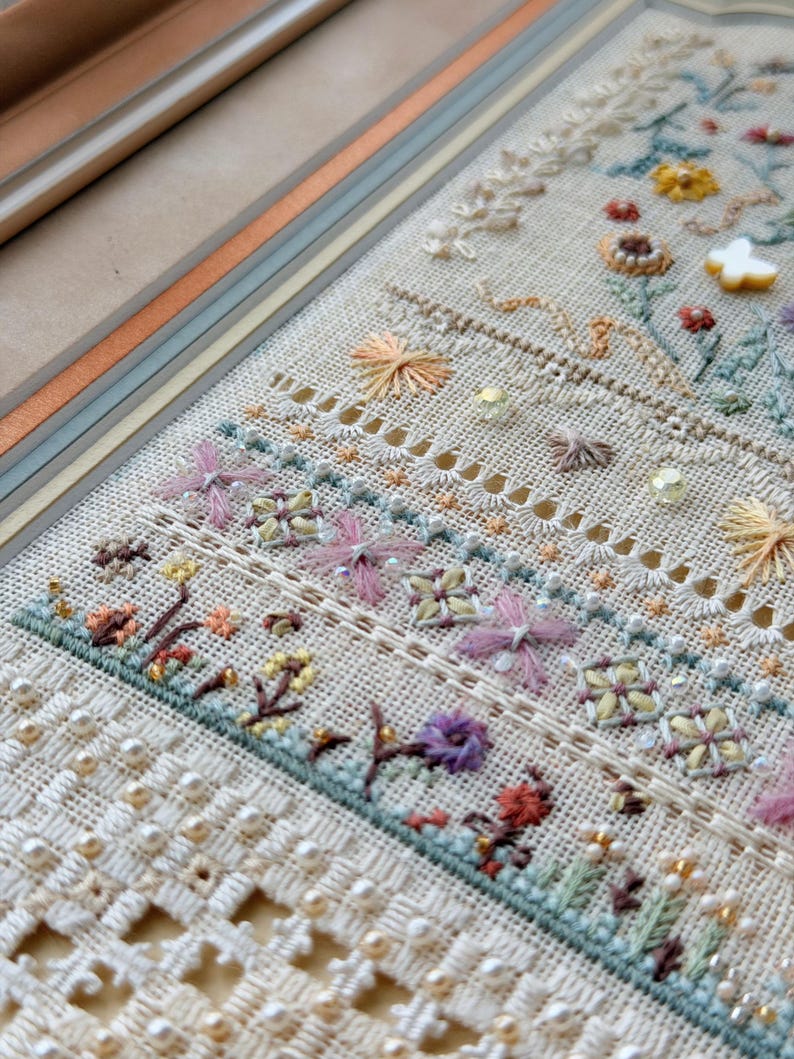 May include: A sampler of intricate needlework on light beige fabric featuring horizontal bands of floral embroidery, drawn thread work, and decorative stitching. The design includes small beads, a white butterfly charm, and pastel-colored threads.