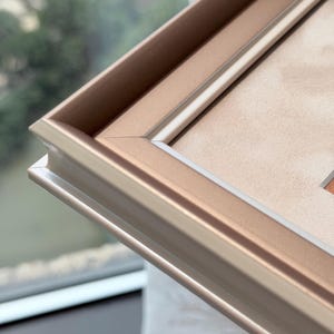 May include: A metallic rose gold picture frame featuring a stepped, multi-layered profile with clean mitered corners. The frame has a smooth, satin-like finish and surrounds a light beige mat board, creating a structured border for artwork.