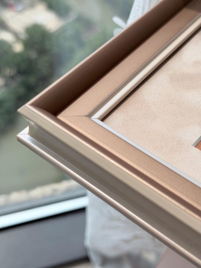 May include: A metallic rose gold picture frame featuring a stepped, multi-layered profile with clean mitered corners. The frame has a smooth, satin-like finish and surrounds a light beige mat board, creating a structured border for artwork.