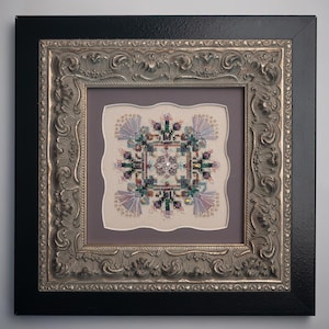 May include: Framed cross-stitch artwork featuring a symmetrical floral design with purple, green, and silver beads. The artwork is set against a light background and framed in a silver ornate frame with a black outer frame.