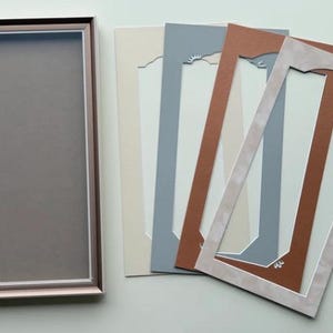 May include: A rectangular picture frame with a metallic rose gold finish and a dark gray backing, accompanied by four rectangular mat boards in cream, slate blue, terracotta, and a light beige with a decorative arched cutout design.