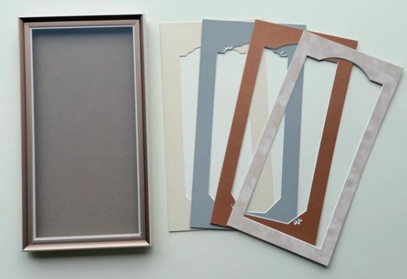 May include: A rectangular picture frame with a metallic rose gold finish and a dark gray backing, accompanied by four rectangular mat boards in cream, slate blue, terracotta, and a light beige with a decorative arched cutout design.
