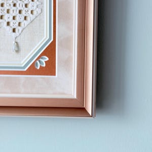 May include: A framed piece of needlework featuring a white macrame-style geometric pattern with small beads, set against a beige fabric background. The frame is a metallic rose gold color with a multi-layered matting in white, blue, and rust orange.