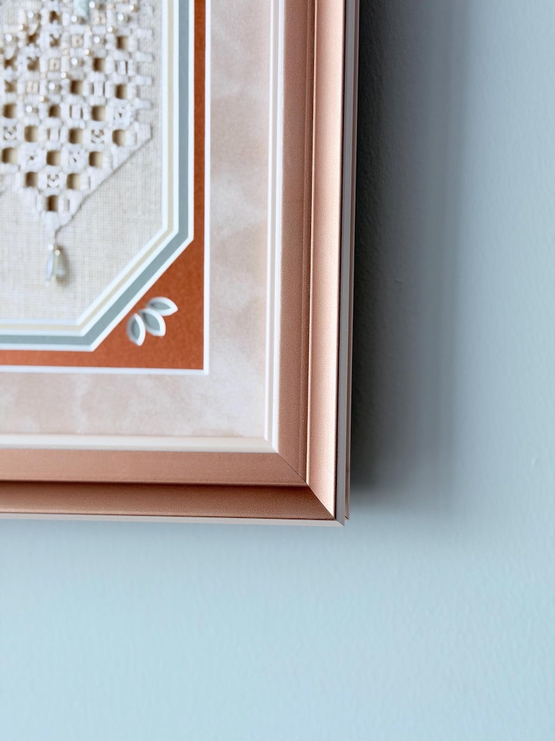May include: A framed piece of needlework featuring a white macrame-style geometric pattern with small beads, set against a beige fabric background. The frame is a metallic rose gold color with a multi-layered matting in white, blue, and rust orange.