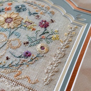 May include: A framed embroidery piece on beige linen fabric featuring a floral arrangement with silk ribbon flowers, intricate cross-stitch botanical motifs, and small beads. The artwork is encased in a multi-layered matting with scalloped edges.