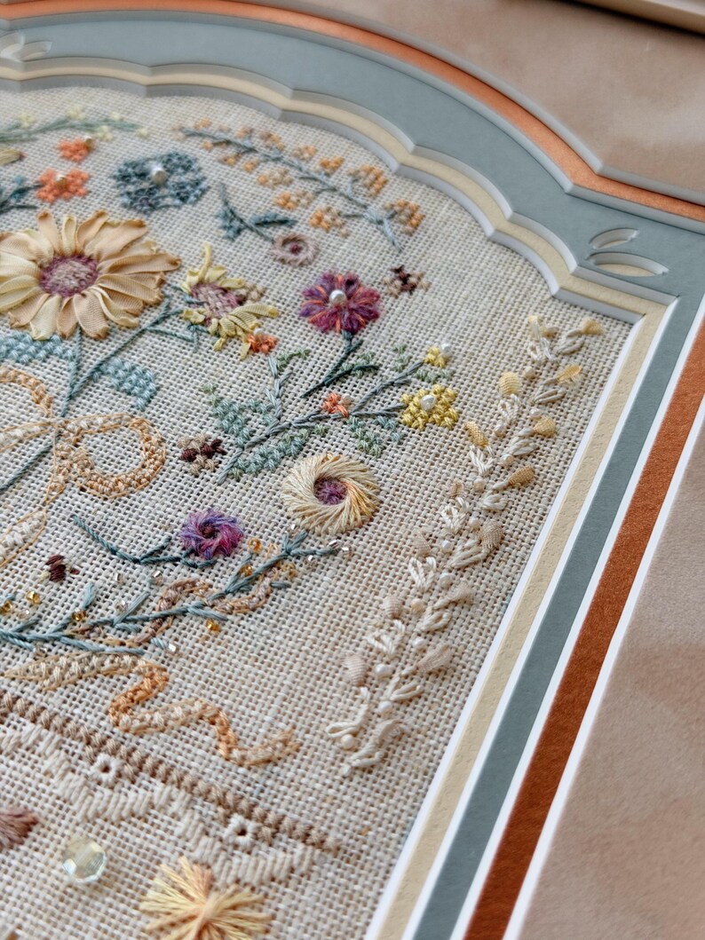 May include: A framed embroidery piece on beige linen fabric featuring a floral arrangement with silk ribbon flowers, intricate cross-stitch botanical motifs, and small beads. The artwork is encased in a multi-layered matting with scalloped edges.