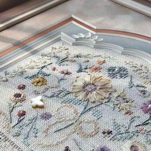 May include: A framed embroidery piece on light-colored fabric featuring a variety of stitched floral motifs in muted tones of yellow, blue, green, and purple. The design includes a central ribbon-style flower, small beads, and a white butterfly charm.