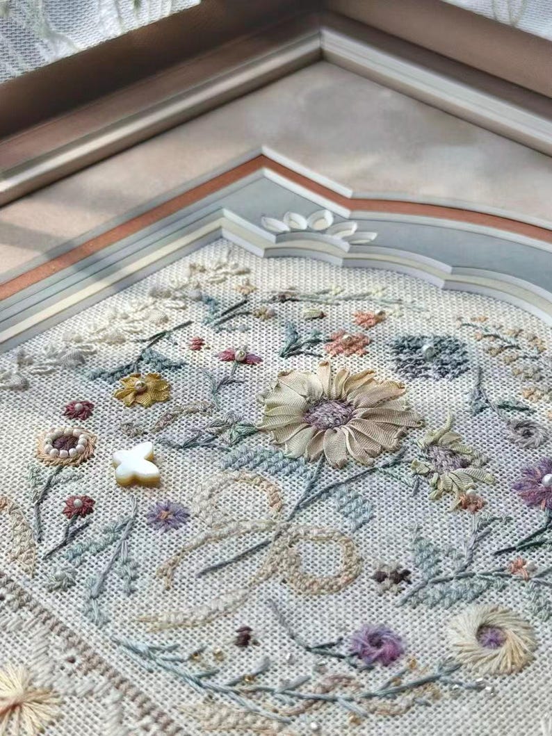 May include: A framed embroidery piece on light-colored fabric featuring a variety of stitched floral motifs in muted tones of yellow, blue, green, and purple. The design includes a central ribbon-style flower, small beads, and a white butterfly charm.