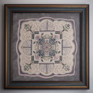 May include: Framed cross-stitch artwork featuring a detailed, symmetrical design in shades of beige, blue, pink, and green. The artwork is framed in a dark blue frame with gold accents, and a decorative border.