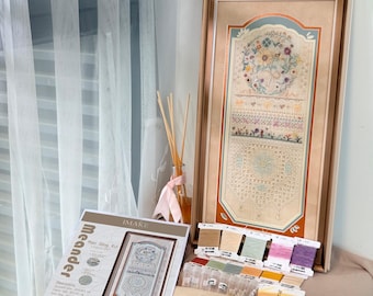 May include: A framed cross-stitch sampler featuring intricate floral and geometric patterns in soft pastel tones, accompanied by a printed instruction sheet labeled Meander, assorted embroidery thread spools, and a piece of beige fabric.