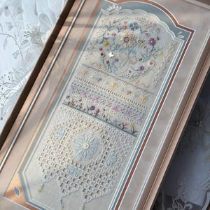 May include: A rectangular framed embroidery sampler featuring intricate needlework with floral motifs, a central butterfly, and geometric lace-like patterns in soft pastel shades of blue, pink, and cream, accented with small beads and crystals.