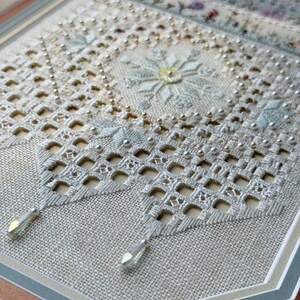 May include: A piece of needlework on light beige linen featuring intricate white cutwork embroidery, a central snowflake motif in pale blue and yellow, small silver beads, and two clear, teardrop-shaped crystal pendants hanging from the bottom.