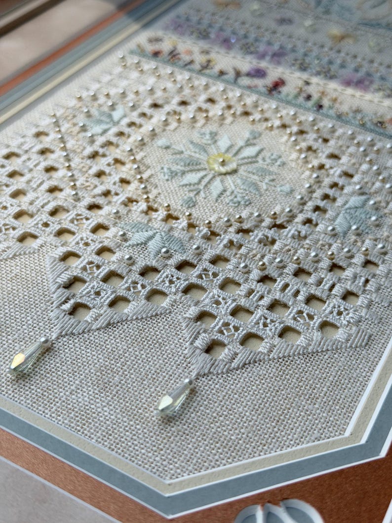 May include: A piece of needlework on light beige linen featuring intricate white cutwork embroidery, a central snowflake motif in pale blue and yellow, small silver beads, and two clear, teardrop-shaped crystal pendants hanging from the bottom.
