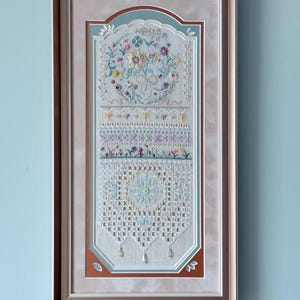 Puede incluir: Una pieza de bordado enmarcada de forma rectangular que presenta una composición vertical de motivos florales, encaje intrincado y patrones geométricos en tonos pastel de azul, rosa y amarillo, terminada con pequeñas cuentas colgantes.