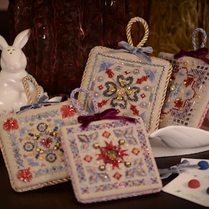 Può includere: Quattro ornamenti quadrati in tessuto beige con ricami complessi in filo rosso, blu e oro, impreziositi da perline e paillettes. Ogni ornamento presenta un cappio in corda intrecciata per appenderlo e un piccolo fiocco in velluto.
