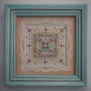 May include: A framed cross-stitch artwork featuring a central floral design in pastel colors. The artwork is surrounded by a light pink mat with a scalloped edge, and a teal-colored wooden frame. The cross-stitch includes beads and intricate stitching.