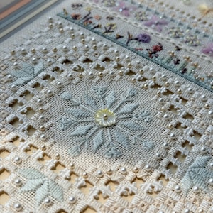 May include: A piece of cream-colored fabric featuring intricate needlework, including cutwork embroidery, light blue floral stitching, and small white beads. A central flower motif is accented with a small, iridescent mother-of-pearl button.