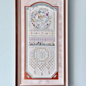 May include: A framed embroidery piece featuring a vertical composition of intricate floral motifs, geometric patterns, and lace-like needlework in soft pastel tones. The artwork is encased in a metallic rose gold frame with a multi-layered matting.