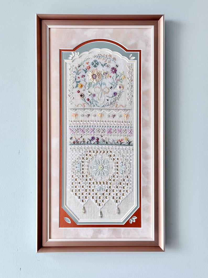 May include: A framed embroidery piece featuring a vertical composition of intricate floral motifs, geometric patterns, and lace-like needlework in soft pastel tones. The artwork is encased in a metallic rose gold frame with a multi-layered matting.