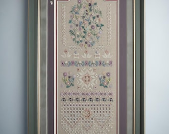 May include: A framed sampler featuring intricate embroidery on a light beige linen fabric. The design includes a floral bouquet at the top, geometric patterns, and a lace-like border at the bottom, all enclosed in a dark green frame with a purple mat.