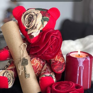 Könnte beinhalten: Ein Teddybär aus rotem Stoff mit beigem und rotem Blumenmuster, der eine mit Bindfaden und einem Metallschlüssel-Anhänger versehene braune Papierrolle hält. Daneben stehen eine rote Stumpenkerze und eine rote Samtrose, mit dem Text A HUG IN A BOX FOR HER.