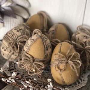 May include: A group of Easter eggs made from tan fabric, some printed with musical notation. Each egg is wrapped with twine and tied with a bow. The eggs are displayed on a white lace doily with white floral decorations.