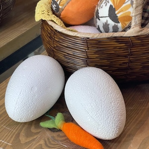 Könnte beinhalten: Zwei große weiße, strukturierte Schaumstoffeier und eine orangefarbene Filzkarotte mit grünen Blättern liegen auf einer Holzoberfläche. Ein geflochtener Korb enthält dekorative Stoffeier mit Blumenmustern.